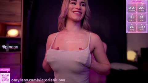 Video of a smiling woman with light skin and blonde hair, wearing a revealing white tank top, in a dimly lit room with purple lighting.