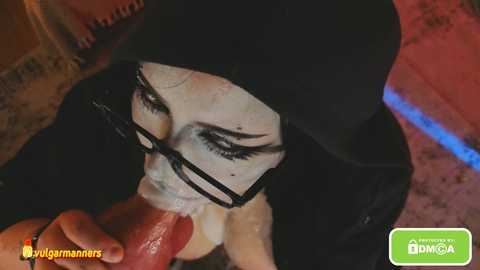 Video of a woman with black eye makeup, wearing a black hooded cloak, performing oral sex on a man with a large erect penis, in a dimly lit room.