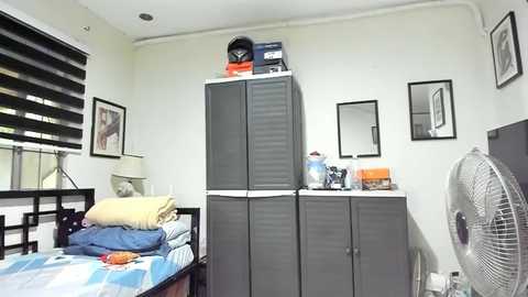 Video of a cluttered, minimalist bedroom with a bed, grey wardrobe, white fan, framed artwork, and blinds; neatly stacked clothes on top of wardrobe.