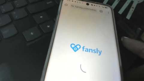 A video of a person's hand holding a smartphone displaying the FanSly app icon on a black keyboard. The background shows a blurred, dark-colored surface.