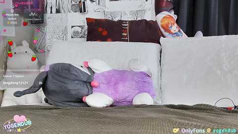 Video of a plush grey cat with a pink nose on a white couch, surrounded by pillows, with a poster and a stuffed animal in the background.