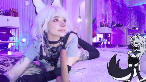 Video of a white-skinned, light-haired, anime-style cosplayer with fox ears and tail, wearing a gray graphic tee and black lace, lying on a bed. The room has purple lighting, shelves, and a small dog toy. Overlay: anime-style black wolf character.