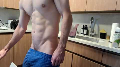 Video of a shirtless, muscular man in blue shorts, standing in a modern kitchen with wooden cabinets, a sink, and various appliances.