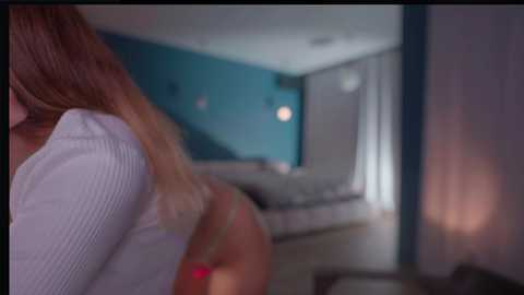 Media: A blurred video of a woman in a white top, with long blonde hair, in a dimly lit, modern bedroom with teal walls and a bed.