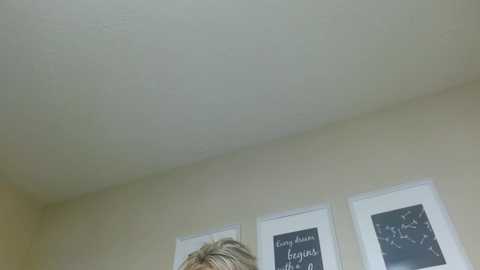 A video of a ceiling with a slight crack, featuring two framed artworks on beige walls, one with a blackboard-like texture and the other with white text. A person with short, blonde hair is partially visible in the foreground.