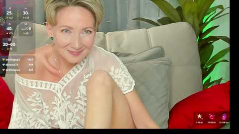 Video of a blonde woman with fair skin and green eyes, wearing a sheer white lace dress, sitting on a red couch, smiling.