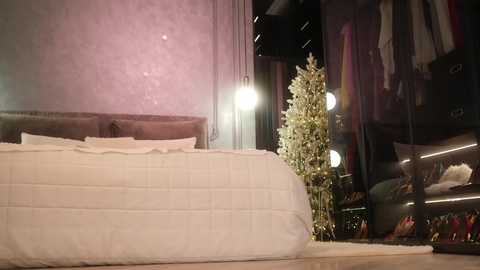 Media: Video of a modern bedroom with a plush, white quilted bed, soft pink wallpaper, and a tall, green floral arrangement by a window.