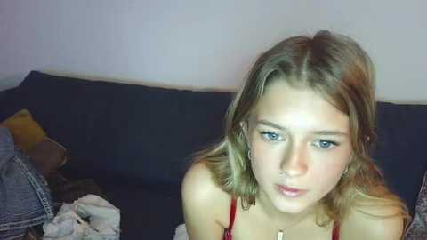 A video of a young woman with fair skin, light brown hair, and blue eyes, wearing a red top, sitting on a dark couch.
