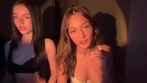 Video of two young women with fair skin and straight, shoulder-length hair, one in a black bra, the other in a beige bra, against a dimly lit background.