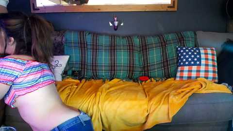 Media: Video of a woman in a striped top, leaning against a plaid couch with a yellow blanket, surrounded by plaid and checkered pillows, in a cozy, dimly lit room.