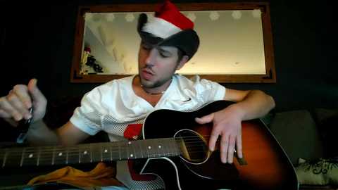 A video of a man in a white shirt and Santa hat, playing an acoustic guitar in a dimly lit room with a mirror and a brown couch.