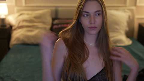 Video of a young woman with long, straight brown hair, wearing a black sleeveless top, sitting on a bed with green sheets, in a dimly lit bedroom with white walls and a nightstand with a lamp.