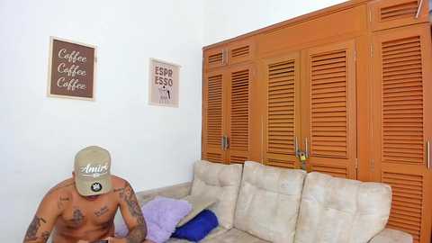 Video of a shirtless man with tattoos, wearing a beige cap, sitting on a beige sofa with a purple cushion, in a room with wooden louvered closet doors and \"Espresso Coffee\" wall art.