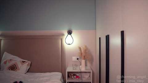 Video of a minimalist bedroom with a beige headboard, a single bedside table holding a vase, and a modern, exposed light bulb wall sconce.