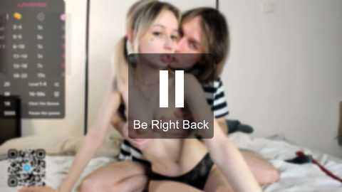 Video of a young couple in a bedroom. The woman, with blonde hair and small breasts, is topless, wearing only black lace panties. The man, with long dark hair, is shirtless, embracing her. Background shows a calendar and a bed with white sheets.