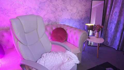 Video of a plush, light gray, tufted armchair with a red pillow, illuminated by soft purple lighting, in a cozy, modern room with white walls, a small table, and a mirror.