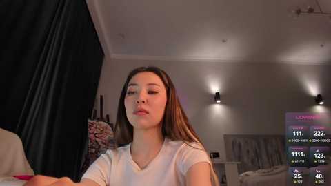 Video of an Asian woman with long black hair, wearing a white t-shirt, seated in a dimly lit bedroom with a TV screen displaying live stream stats in the background.