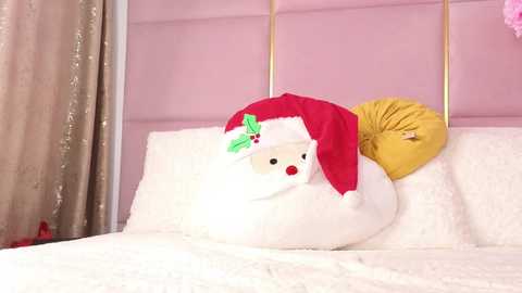 Media: A video of a plush Santa Claus doll wearing a red hat and yellow scarf, sitting on a white bed with pink headboard, surrounded by cream curtains.