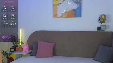 Video of a modern, minimalist bedroom with a beige headboard, pink and gray pillows, a pink bear, a potted plant, and abstract wall art.