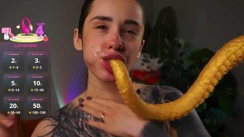 A photo of a young woman with light skin, long dark hair, and a tattoo on her shoulder, licking a large, yellow, cartoonish banana. Background shows a blurry plant and a digital game interface.