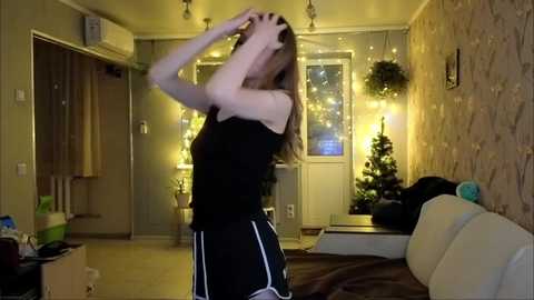 Video of a young woman in a black top and white-striped pajama bottoms, dancing in a cozy, dimly lit bedroom with a Christmas tree and fairy lights.