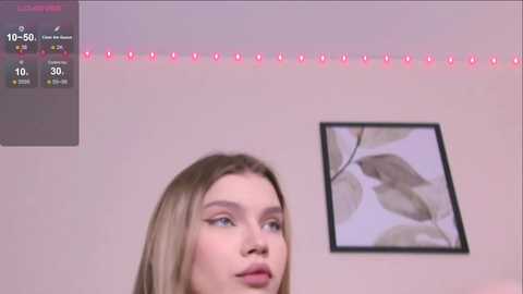 Video of a young woman with fair skin and straight, shoulder-length blonde hair, wearing makeup, standing in front of a wall with a framed leaf pattern painting. A digital display with temperature and humidity readings is visible in the upper left corner.