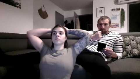 Video of a young woman with tattoos, wearing a gray t-shirt, sitting on a couch in a dimly lit living room, holding her hair. A bearded man in a striped shirt sits beside her.