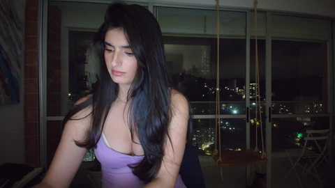 Video of a brunette woman with long hair, wearing a purple strapless dress, standing indoors by a window with a city skyline view at night.