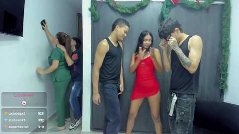 Video of a live stream: Three shirtless Asian men, one with a tattoo, in a red dress, and a woman in green scrubs, laughing.