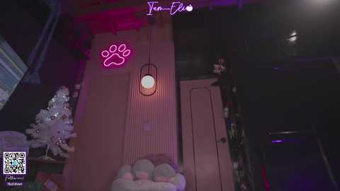 Video of a dimly lit room with a pink paw print neon sign, a small white Christmas tree, and a plush toy on a pink and white striped wall.