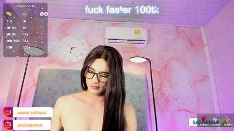 A video of an Asian woman with long black hair, wearing glasses, topless, standing in a pastel-colored room with a clock, air conditioner, and a lamp.