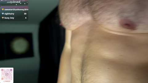 Video of a shirtless man's bare chest, with visible nipples and a faint mole. Background shows a blurry, dimly-lit room with a circular clock and a dark curtain.