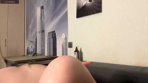 A video of a person lying on a dark couch, with a large skyscraper wallpaper and a black-and-white abstract art print on the wall. The image captures the upper body and head, with a focus on the person's bare, smooth back and shoulders.