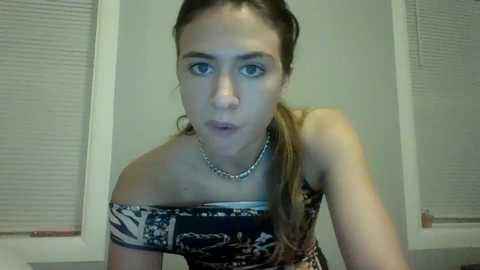 Video of a young woman with light skin and brown hair, wearing a strapless dress with a floral pattern, indoors with white blinds in the background.