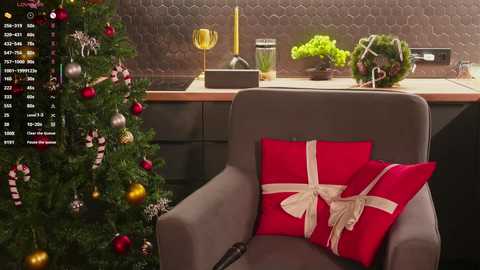 A video of a cozy living room featuring a decorated Christmas tree, a grey armchair with a red pillow and a gift, a wooden kitchen counter, and a modern, geometric-patterned wall.