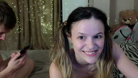 Video of a young, smiling woman with long, ombre hair in pigtails, topless, holding a phone, in a dimly lit room with string lights, stuffed animals, and a zebra-patterned blanket.