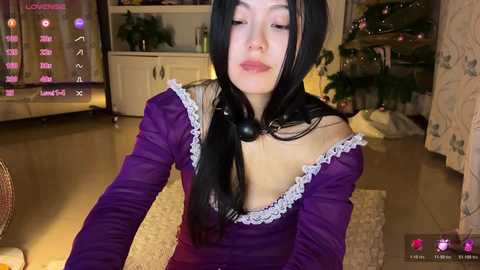 Video of an Asian woman with long black hair, wearing a purple off-shoulder dress with lace trim, indoors near a TV showing a pink screen.