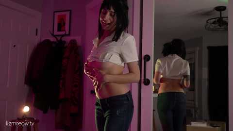Video of a slender, fair-skinned woman with shoulder-length black hair, lifting her white crop top to expose her bare breasts, standing in a dimly-lit hallway with a mirror reflecting her back.