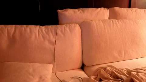 A video of a beige sofa with plush cushions, a black wall in the background, and a beige blanket lying on the floor in front.