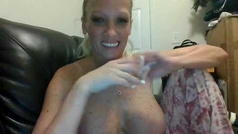 Video of a smiling, topless blonde woman with medium-sized breasts, wearing a floral-patterned dress, sitting on a black leather couch. Background shows a white door and hanging clothes.