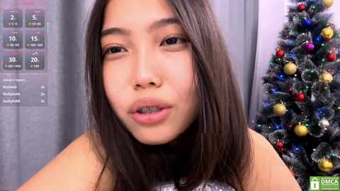 Video of an Asian woman with long black hair and light skin, wearing a gray top, standing in front of a Christmas tree and a weather chart in a room.