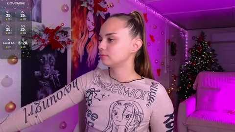 Video of a young woman with light skin and long brown hair in a ponytail, wearing a long-sleeved graphic tee with text and images, standing indoors with festive decorations.