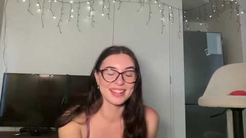 Media: Video of a smiling woman with long dark hair and glasses, sitting indoors in a cozy room with a flat-screen TV, string lights, and a beige armchair.