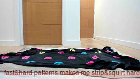 A video of a minimalist room with light wood floors, a black blanket with colorful, cartoon-like patterns, and a beige carpet. The room features a closed wooden door and white walls. Text at the bottom reads, \"fast & hard patterns make me strip & squirt hare.\