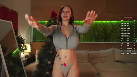 Media: Video of a woman with large breasts, wearing a light blue crop top and matching thong, standing in a modern living room with a Christmas tree and green wall.
