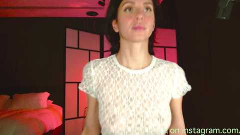 Media: Video of a young woman with fair skin and straight black hair, wearing a white, lace-patterned top, standing in a dimly lit room with red lighting, featuring a black and white shoji screen and a bed in the background.
