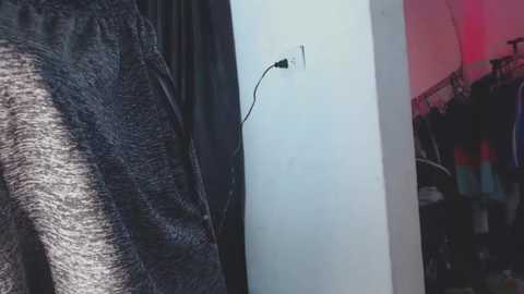 Video of a person wearing dark grey sweatpants with a black electrical cord plugged into a white wall socket. Background features a dark curtain and colorful clothing items.