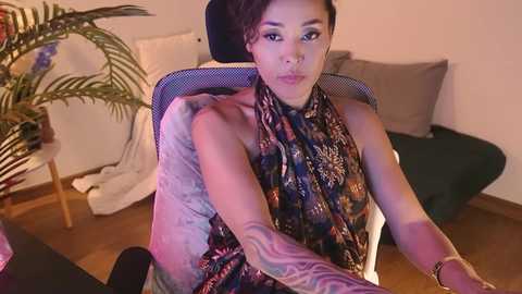 Media: Video of an Asian woman with short black hair, wearing a patterned scarf, sitting in a chair, surrounded by potted plants and cushions in a cozy, dimly-lit room.
