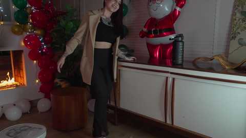Media: Video of a woman in a beige coat and black pants standing near a lit Christmas tree and a large inflatable Santa Claus, in a cozy, festive room.