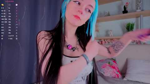 A young woman with long, blue-black hair and tattoos poses in a cozy, floral bedroom. She wears a white lace top and a blue bracelet, smiling slightly.